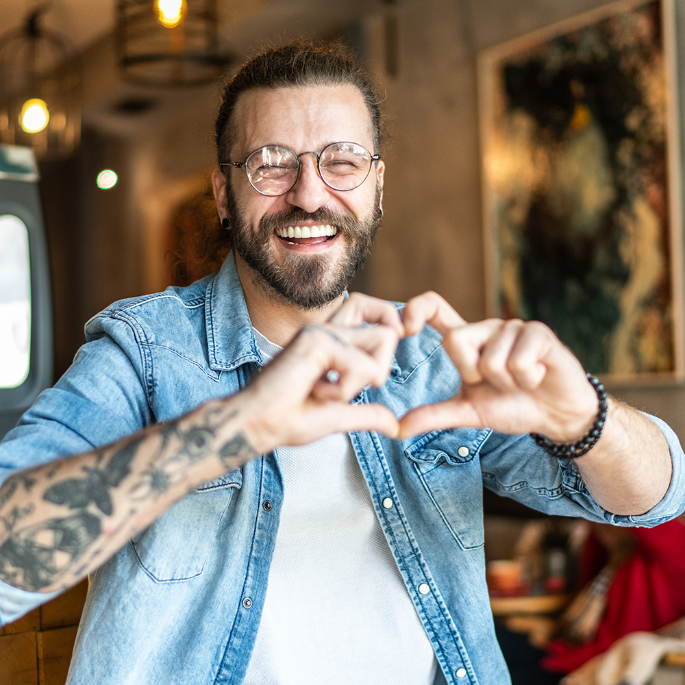 A photo of a man with tattoo sleeves looking directly at the camera and smiling. He's making a heart gesture with his hands.
