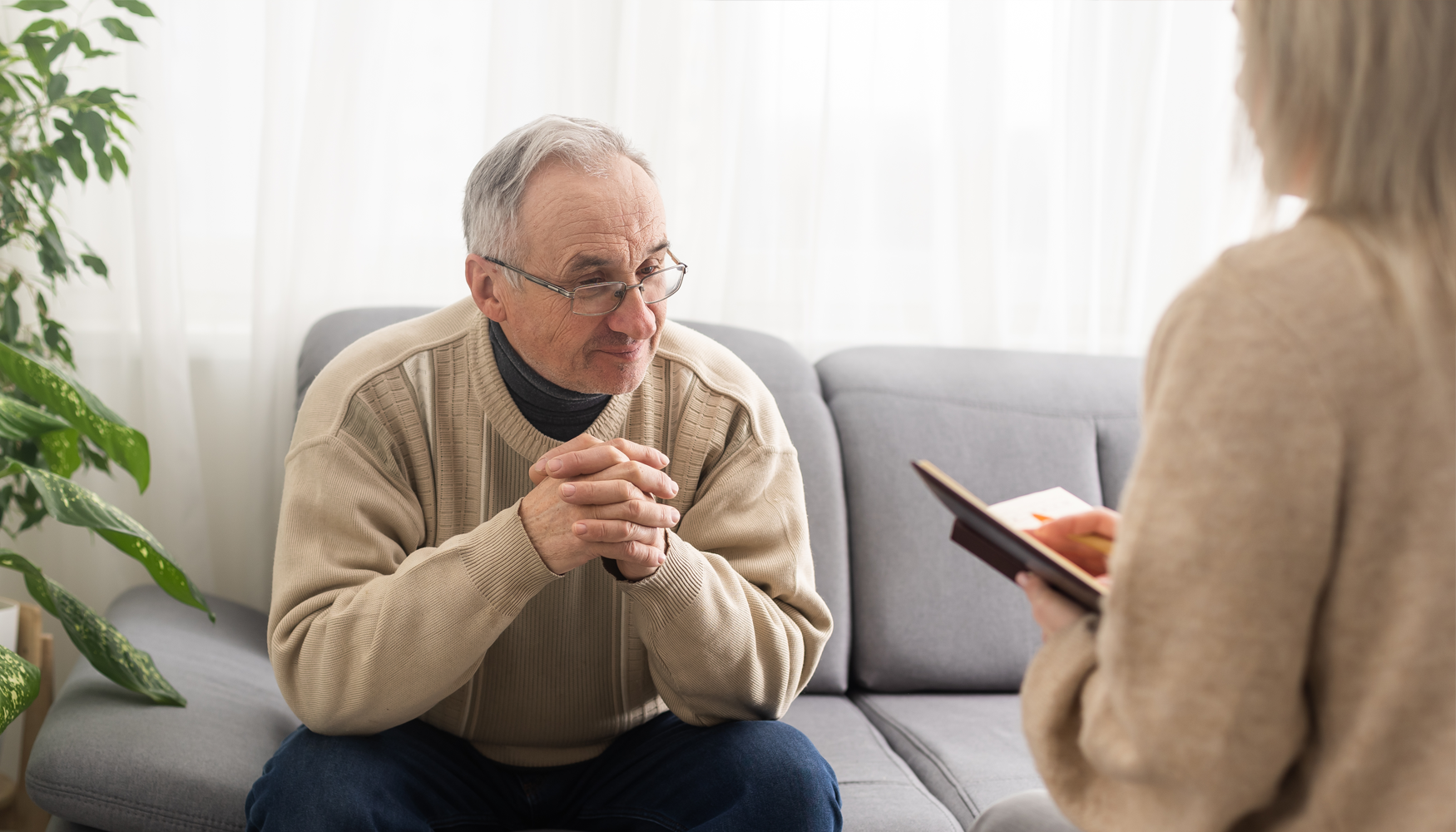 A photo of a senior, caucasian man sitting on a couch listening to his counsellor.